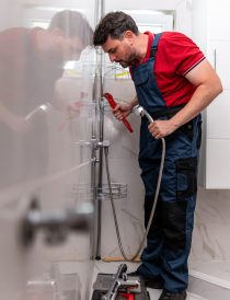 A plumber in a red shirt and blue overalls works on fixing a shower in a clean and bright bathroom.