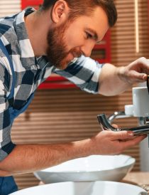 Uses a screwdriver for repairs. Plumber in blue uniform is at work in the bathroom.